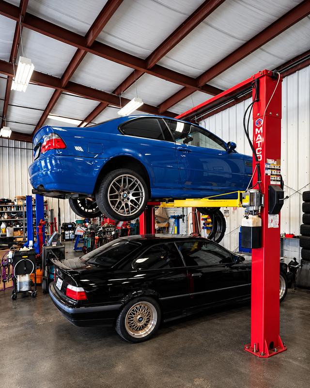 A blue BMW E46 M3 raised on a two-post lift above a black E36 in the German Performance shop